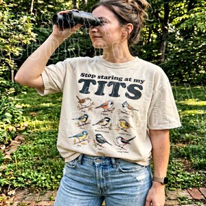 May include: Beige t-shirt with the text "Stop staring at my TITS" in black block letters. Below the text are illustrations of various birds. The person is wearing blue jeans and holding binoculars.