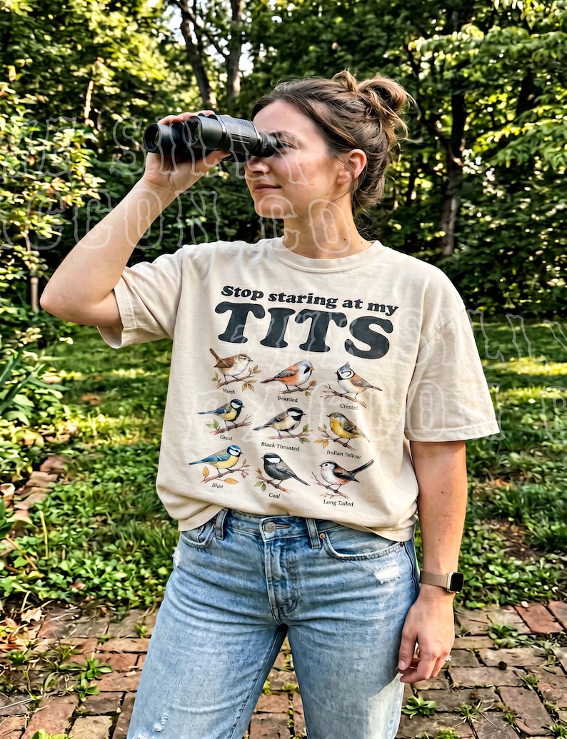 May include: Beige t-shirt with the text "Stop staring at my TITS" in black block letters. Below the text are illustrations of various birds. The person is wearing blue jeans and holding binoculars.