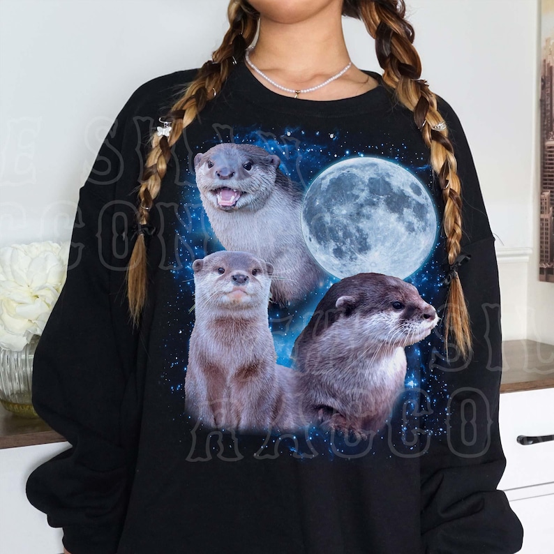 A person wearing a black sweater with a graphic design featuring three otters against a backdrop of a full moon and stars.