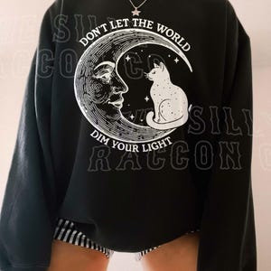 A person wearing a black sweatshirt with a graphic design featuring a cat on a crescent moon and the text "Don't let the world dim your light" printed on it.
