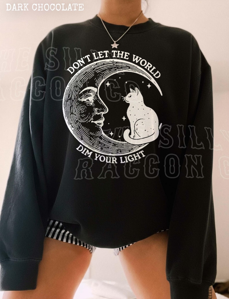A person wearing a black sweatshirt with a graphic design featuring a cat on a crescent moon and the text "Don't let the world dim your light" printed on it.