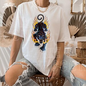 A person wearing a white t-shirt with a graphic design of a black panther with flames on its back, sitting on a chair.