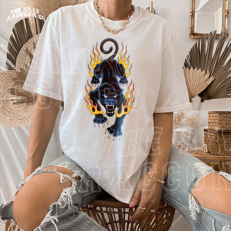 A person wearing a white t-shirt with a graphic design of a black panther with flames on its back, sitting on a chair.