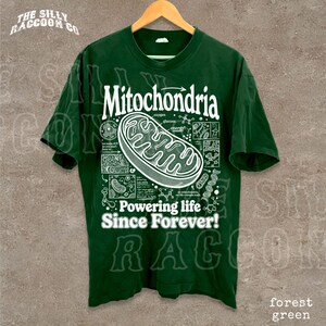 A green t-shirt with a graphic design depicting Mitochondria, a cellular organelle, and text that reads "Mitochondria. Powering life since forever!"