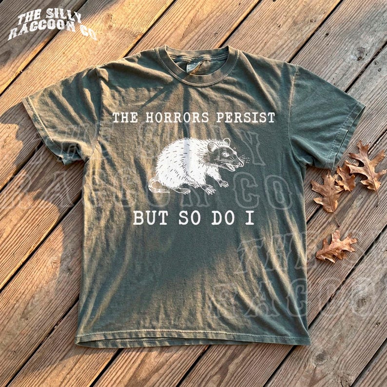 A t-shirt with a humorous text printed on it, lying on a wooden surface with some fallen leaves around it.