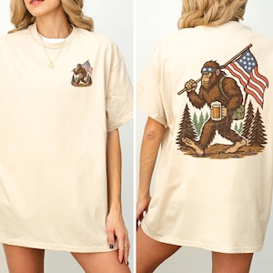 K&ouml;nnte beinhalten: Sandfarbenes T-Shirt mit einem Bigfoot-Motiv und einer amerikanischen Flagge auf der Vorder- und R&uuml;ckseite. Die R&uuml;ckseite zeigt Bigfoot mit Bier und Flagge. Das Shirt hat eine lockere Passform.