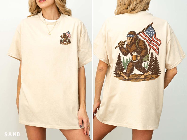 K&ouml;nnte beinhalten: Sandfarbenes T-Shirt mit einem Bigfoot-Motiv und einer amerikanischen Flagge auf der Vorder- und R&uuml;ckseite. Die R&uuml;ckseite zeigt Bigfoot mit Bier und Flagge. Das Shirt hat eine lockere Passform.