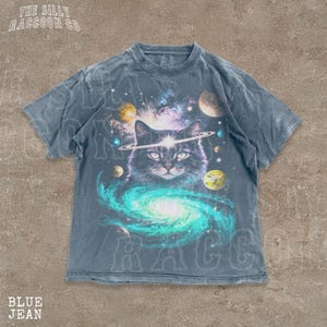 A t-shirt with a graphic design depicting a cat surrounded by a cosmic scene, including planets, stars, and a swirling galaxy.