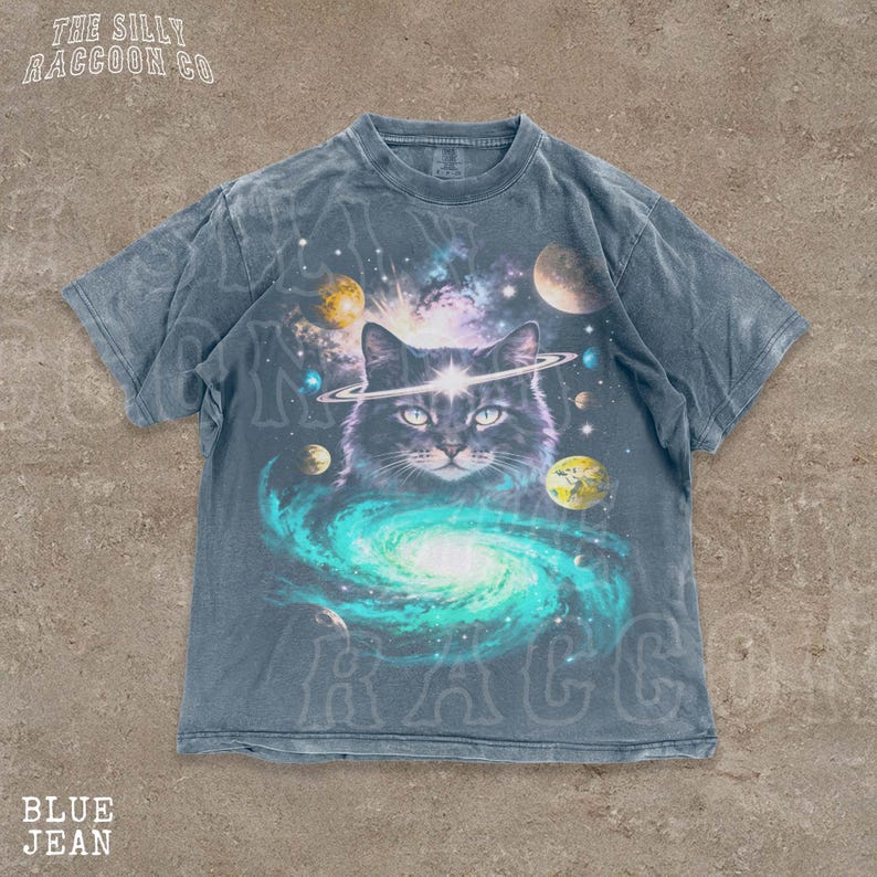 A t-shirt with a graphic design depicting a cat surrounded by a cosmic scene, including planets, stars, and a swirling galaxy.