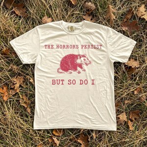 A white t-shirt with a graphic design and text printed on it, lying on a bed of dry grass and leaves.
