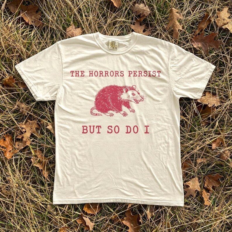 A white t-shirt with a graphic design and text printed on it, lying on a bed of dry grass and leaves.