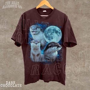 A brown t-shirt with a graphic design featuring three otters against a backdrop of a full moon and blue stars.