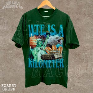 A green t-shirt with a graphic design featuring the Statue of Liberty and an eagle, along with the text "WTE IS A KILOMETER" printed on it.