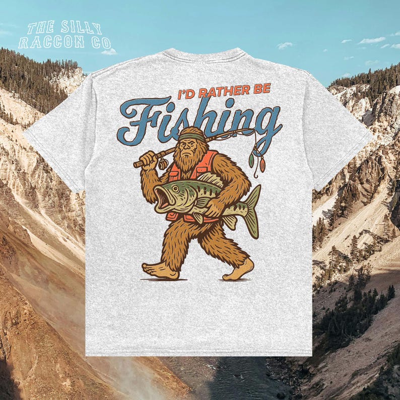 A white t-shirt with a graphic design depicting a large, furry creature holding a fish and the text "I'D Rather Be Fishing" against a mountainous background.