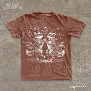 a brown t-shirt with a white illustration of two foxes dancing together, surrounded by stars.
