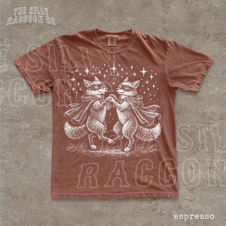 a brown t-shirt with a white illustration of two foxes dancing together, surrounded by stars.