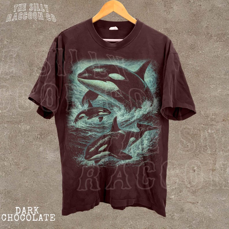 A dark brown t-shirt with a graphic design depicting two orca whales swimming in the ocean.