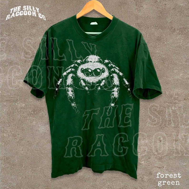A green t-shirt with a white spider graphic printed on it, hanging on a wooden hanger against a concrete wall.