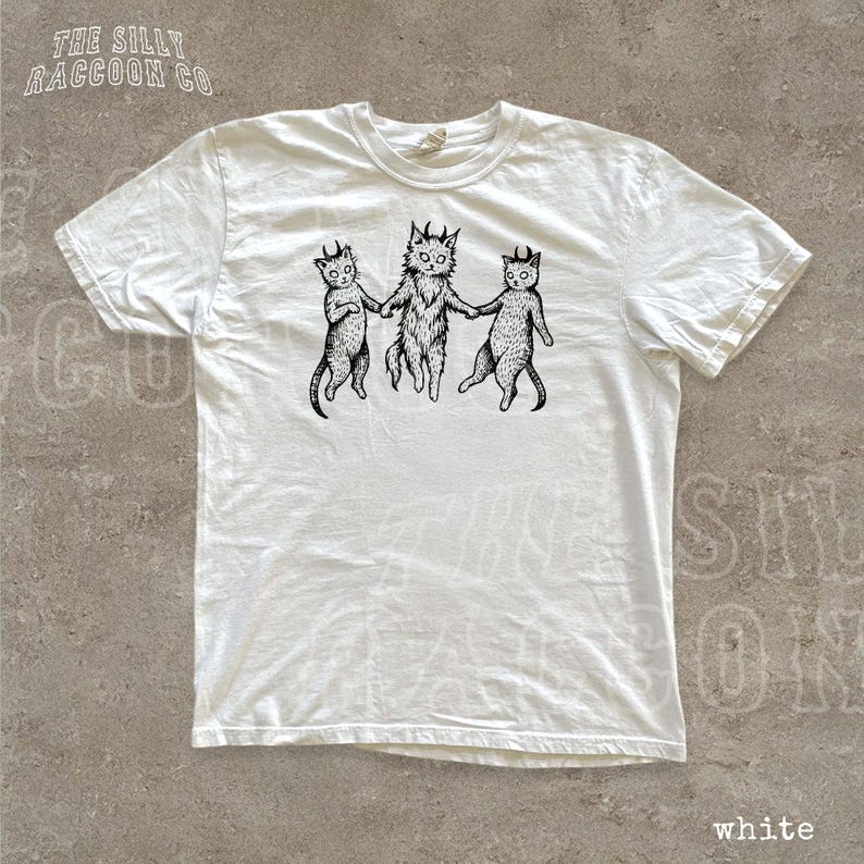 A white t-shirt with a black and white illustration of three cats holding hands.