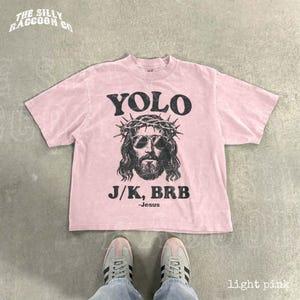A pink t-shirt with a graphic of a man wearing sunglasses and a crown, along with the text "YOLO J.K. BRB Jesus" printed on it. The t-shirt is laid out on a concrete floor, and a pair of sneakers is visible underneath it.