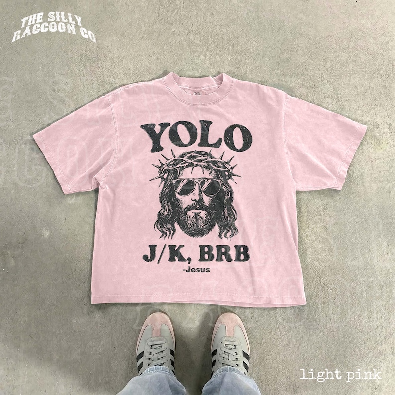 A pink t-shirt with a graphic of a man wearing sunglasses and a crown, along with the text "YOLO J.K. BRB Jesus" printed on it. The t-shirt is laid out on a concrete floor, and a pair of sneakers is visible underneath it.