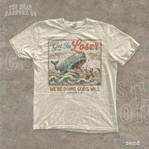A white t-shirt with a graphic design depicting a person being attacked by a large whale, with the text "Get In Loser" and "We're Doing God's Will" printed on it.
