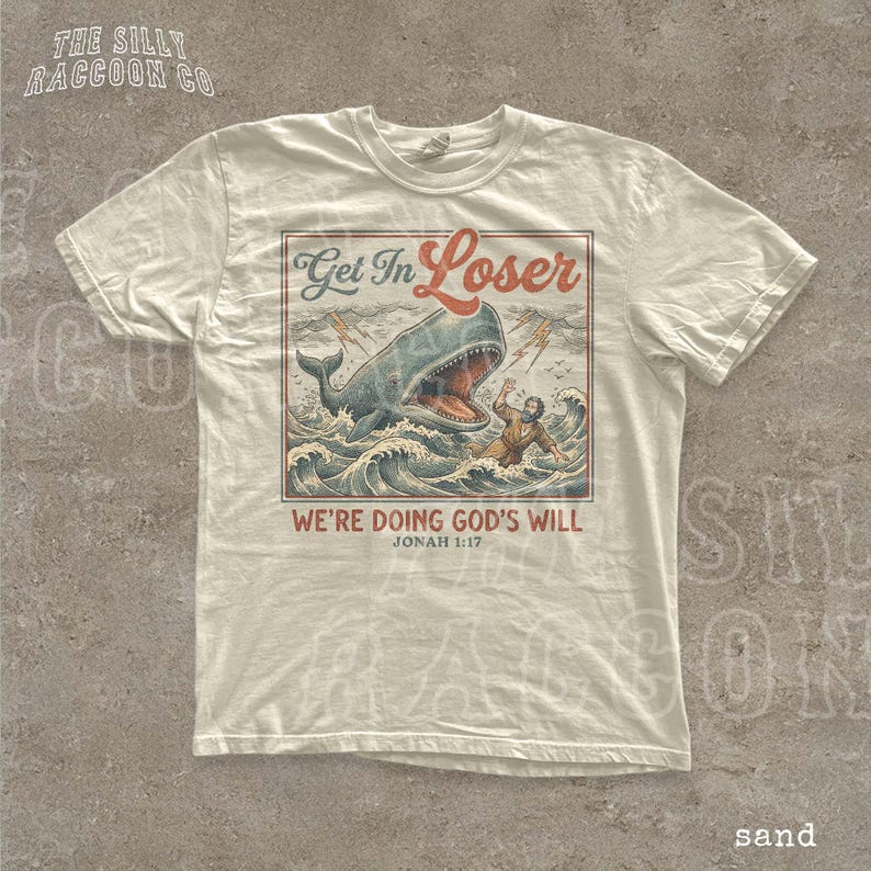 A white t-shirt with a graphic design depicting a person being attacked by a large whale, with the text "Get In Loser" and "We're Doing God's Will" printed on it.