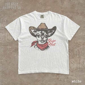 A white t-shirt with a graphic design of a dog wearing a cowboy hat and a red scarf, along with the text "Raise Hell" printed on it.
