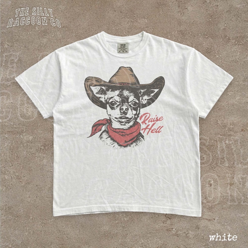 A white t-shirt with a graphic design of a dog wearing a cowboy hat and a red scarf, along with the text "Raise Hell" printed on it.