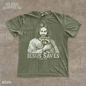 A t-shirt with a graphic design depicting a man holding a raccoon, along with the text "JESUS SAVES" and " Luke 19:10".