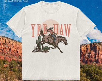 May include: Off-white t-shirt with a western design. The graphic shows a cowboy riding a horse, the words "Yee Haw", a cactus, and a skull. The brand name "The Silly Raccoon Co" is printed at the top left.