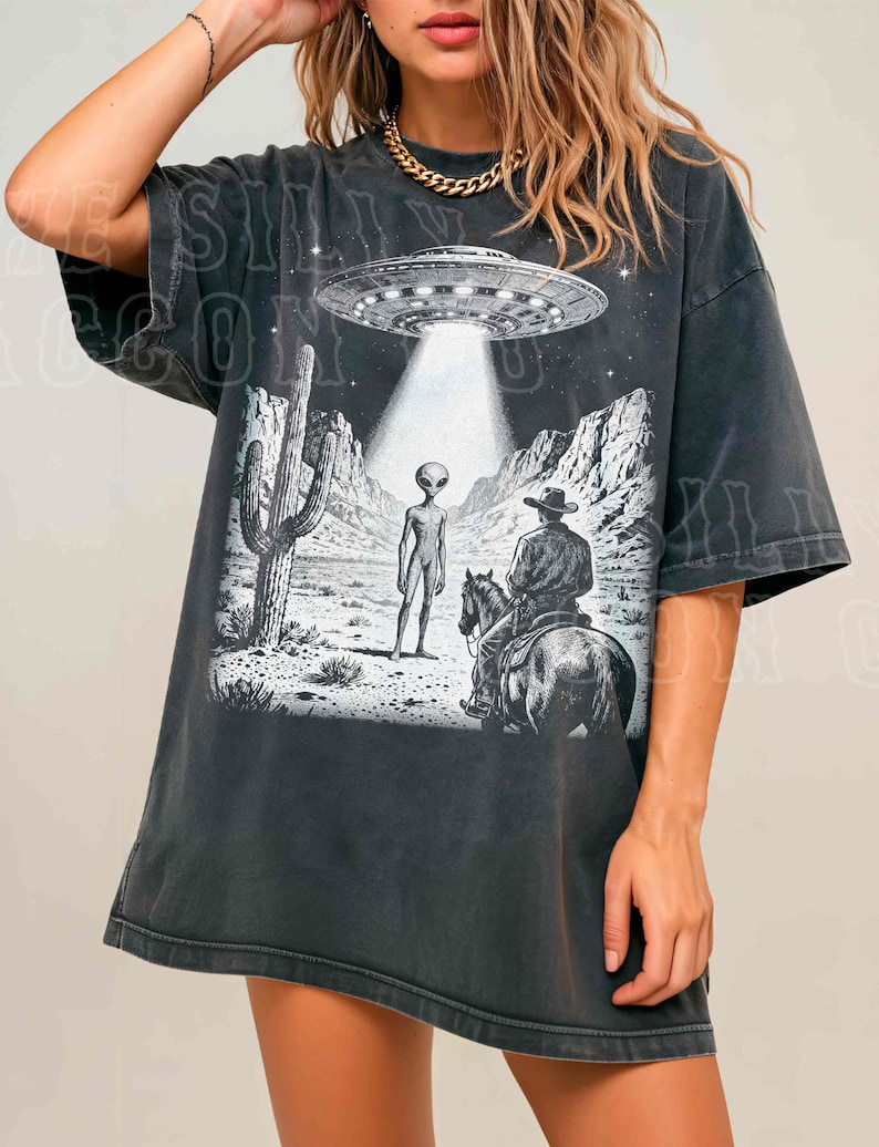 A woman wearing a black t-shirt with a graphic design depicting an alien spacecraft hovering over a desert landscape with a cowboy on horseback.