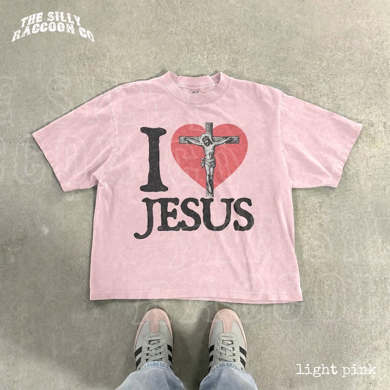 A pink t-shirt with a graphic of a cross and the text "I ♥ JESUS" printed on it, along with a heart symbol. The t-shirt is laid out on a concrete floor, and a pair of sneakers is visible underneath it.