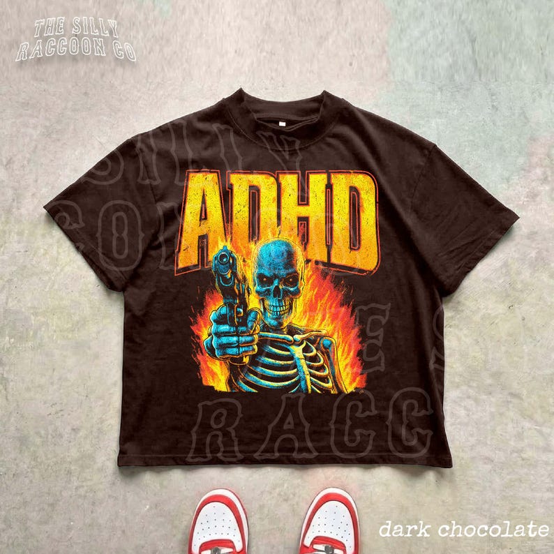 a black t-shirt with a graphic design depicting a skeleton holding a gun, surrounded by flames. The t-shirt is placed on a gray surface, and there are a pair of red and white sneakers visible in the bottom right corner of the image.