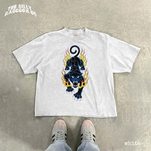 A white t-shirt with a graphic design of a black panther with flames on its back, along with a pair of white sneakers.