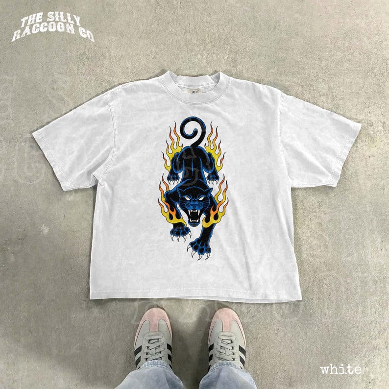 A white t-shirt with a graphic design of a black panther with flames on its back, along with a pair of white sneakers.