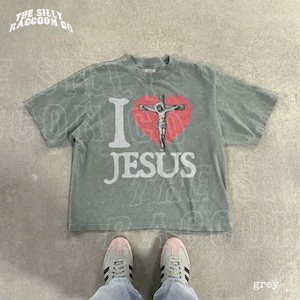 A gray t-shirt with a graphic of a cross and the text "I I heart Jesus" printed on it, along with a red heart symbol. The t-shirt is laid out on a concrete floor, and a pair of white sneakers is visible underneath it.