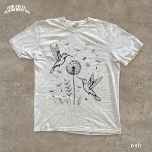 A white t-shirt with a black and white illustration of two hummingbirds hovering over a dandelion and some plants.