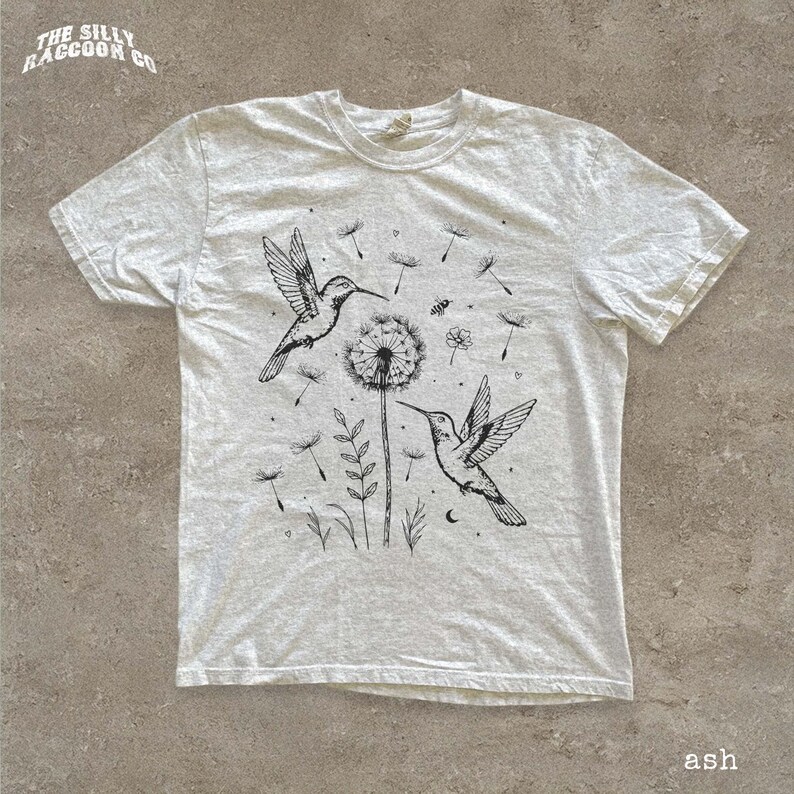 A white t-shirt with a black and white illustration of two hummingbirds hovering over a dandelion and some plants.