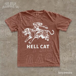 A brown t-shirt with a graphic design depicting a person riding a tiger, accompanied by the text "HELL CAT" and "espresso".