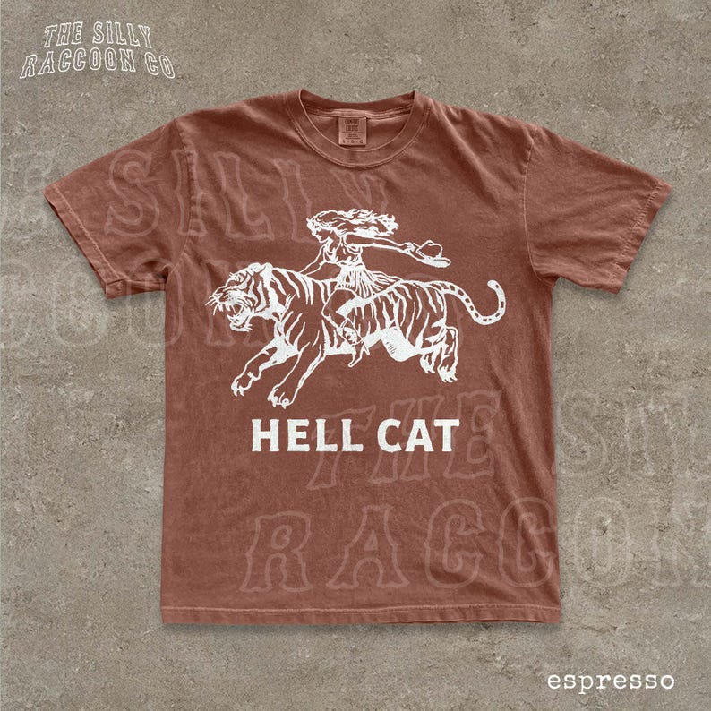 A brown t-shirt with a graphic design depicting a person riding a tiger, accompanied by the text "HELL CAT" and "espresso".