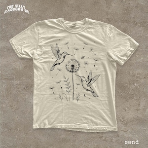 A white t-shirt with a black and white illustration of two hummingbirds feeding on a dandelion, surrounded by other flowers and plants.