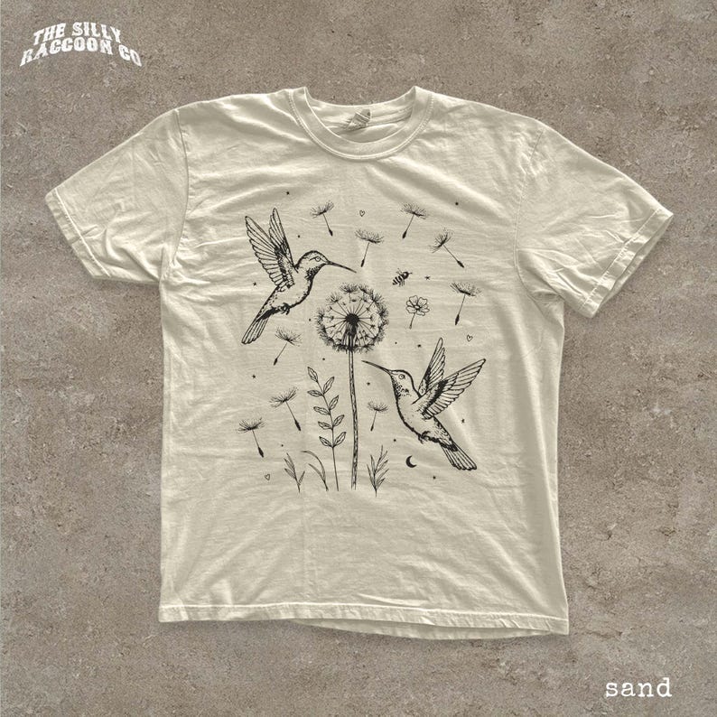 A white t-shirt with a black and white illustration of two hummingbirds feeding on a dandelion, surrounded by other flowers and plants.