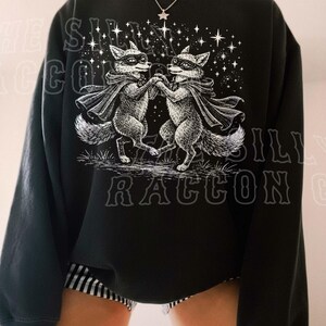 A person wearing a black sweatshirt with a graphic design featuring two raccoons dancing under a starry sky.