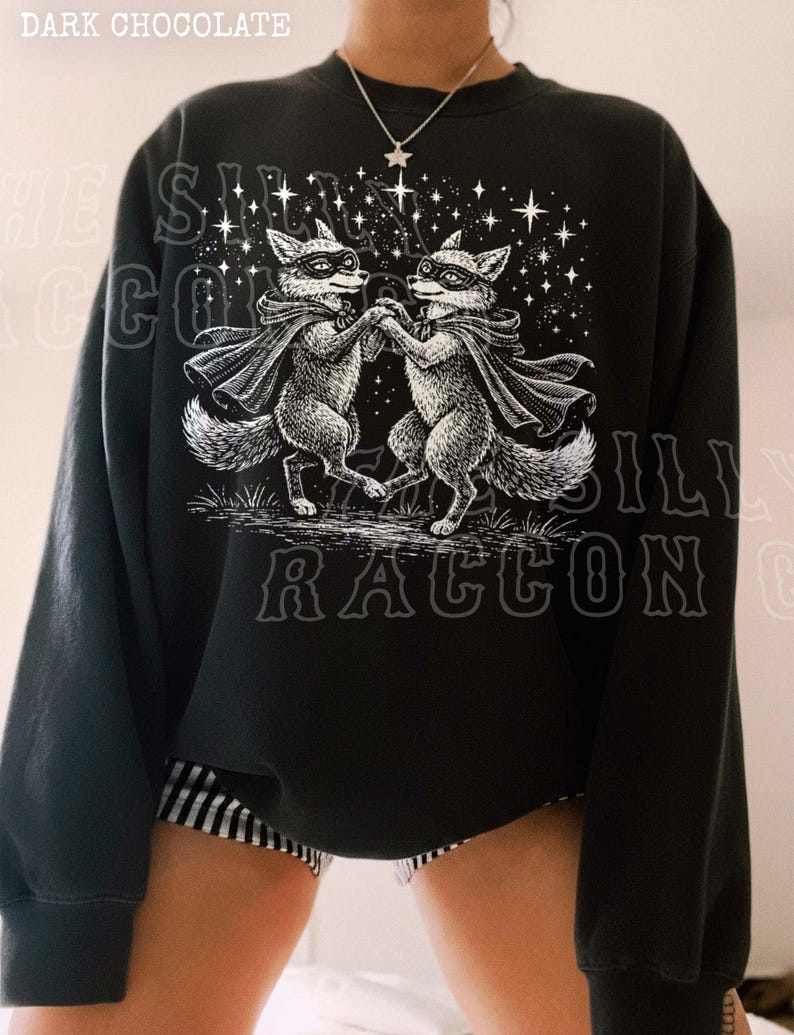 A person wearing a black sweatshirt with a graphic design featuring two raccoons dancing under a starry sky.