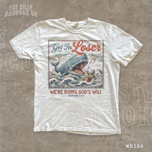 A white t-shirt with a graphic design depicting a man swimming in front of a large whale, with the text "Get In Loser" and "We're Doing God's Will" below the image.
