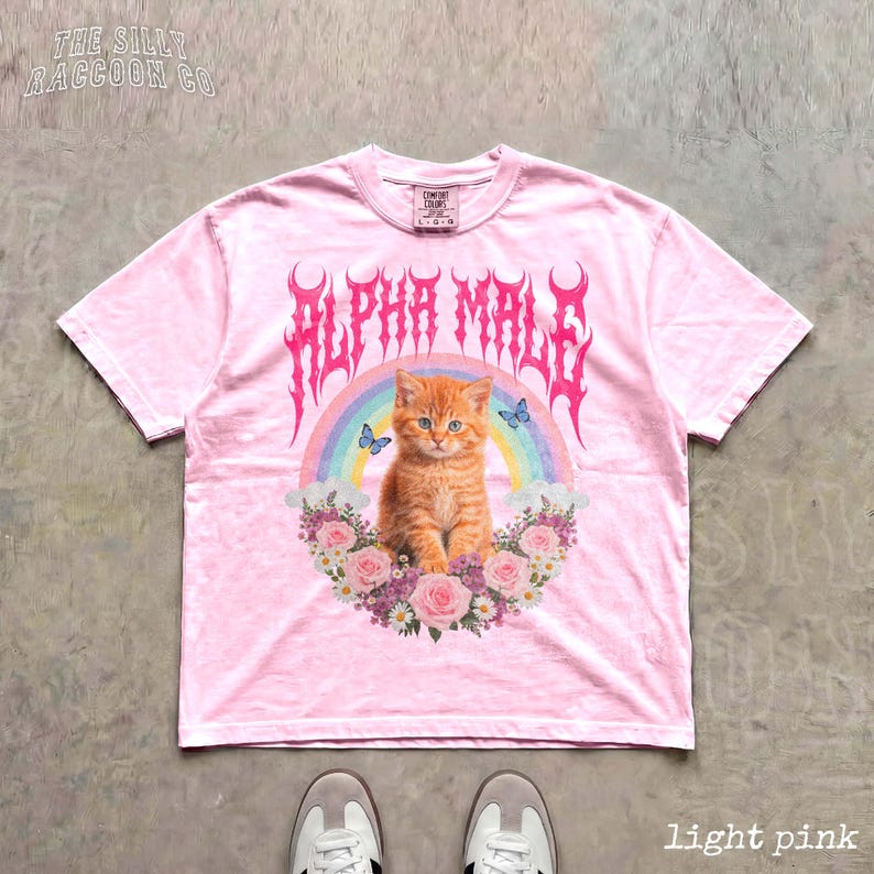 A pink t-shirt with a graphic design featuring an orange cat, a rainbow, and flowers.