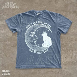 A blue t-shirt with a graphic design depicting a cat sitting on a crescent moon and the text "Don't Let the World Dim Your Light" printed on it.