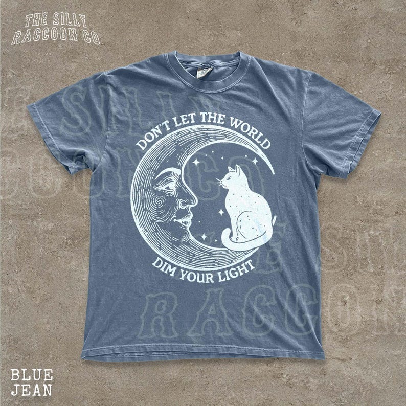 A blue t-shirt with a graphic design depicting a cat sitting on a crescent moon and the text "Don't Let the World Dim Your Light" printed on it.