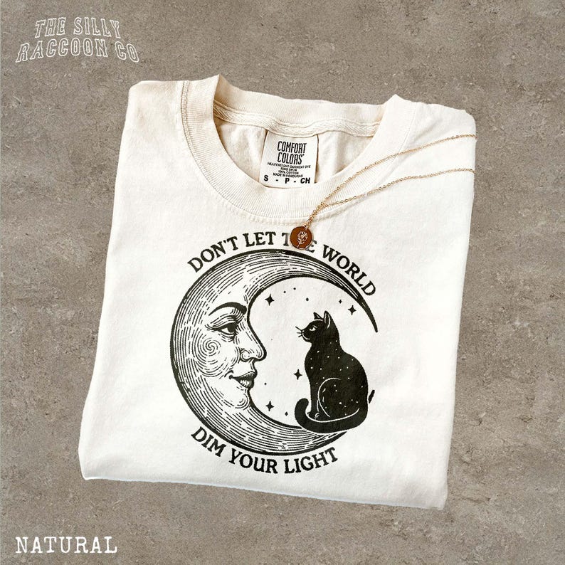 A white t-shirt with a graphic design featuring a cat and a crescent moon, along with the text "Don't let the world dim your light".